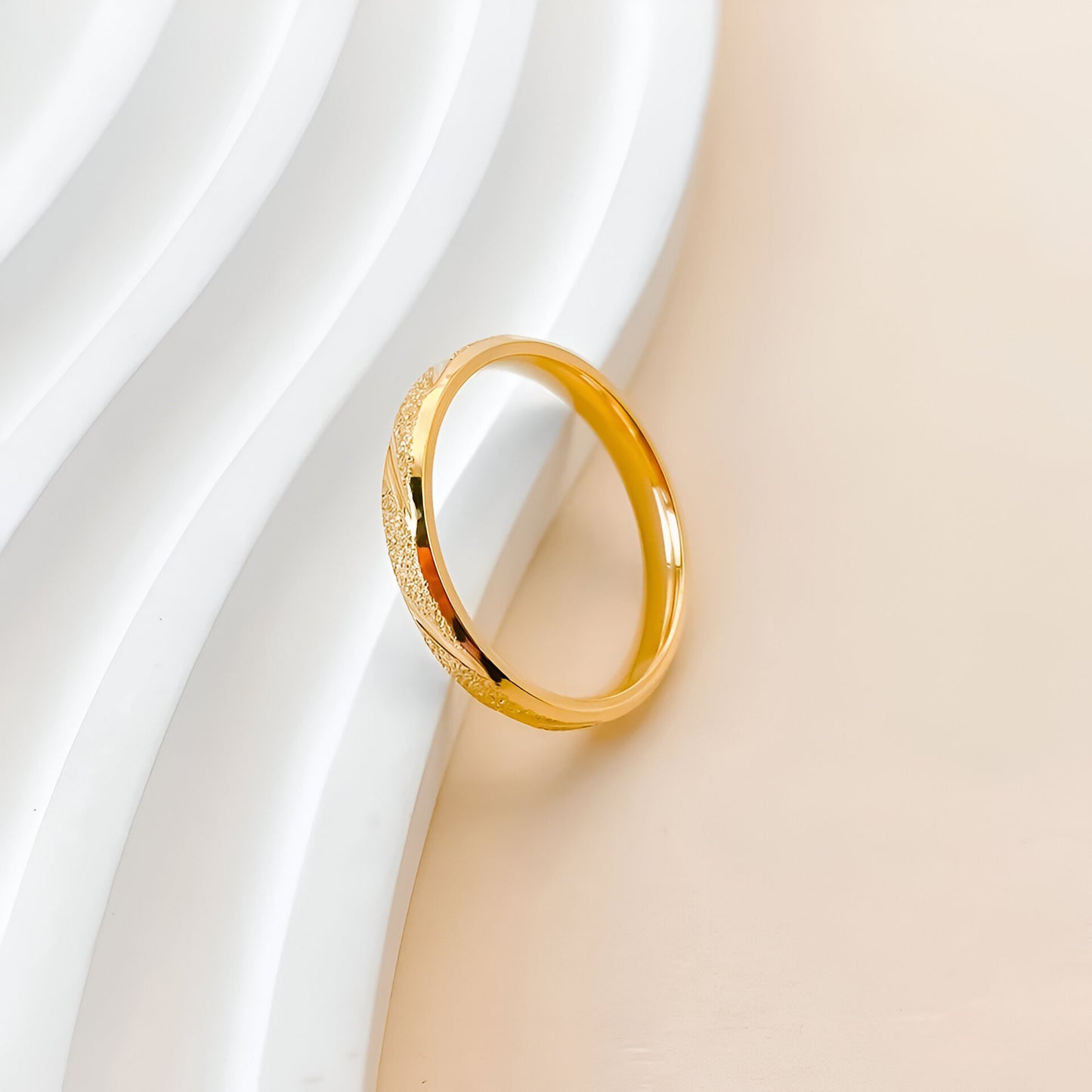 Vue inclinée d’une alliance en or rayée sablée, en acier inoxydable plaqué or 14K, posée sur fond texturé