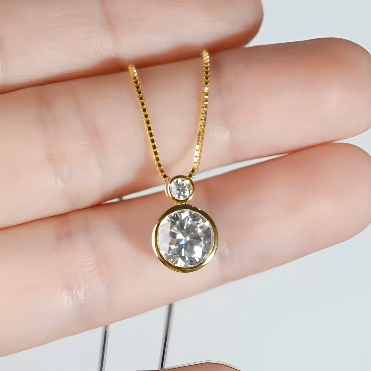 Pendentif serti clos en or jaune et diamants synthétiques entre les doigts d'une femme.