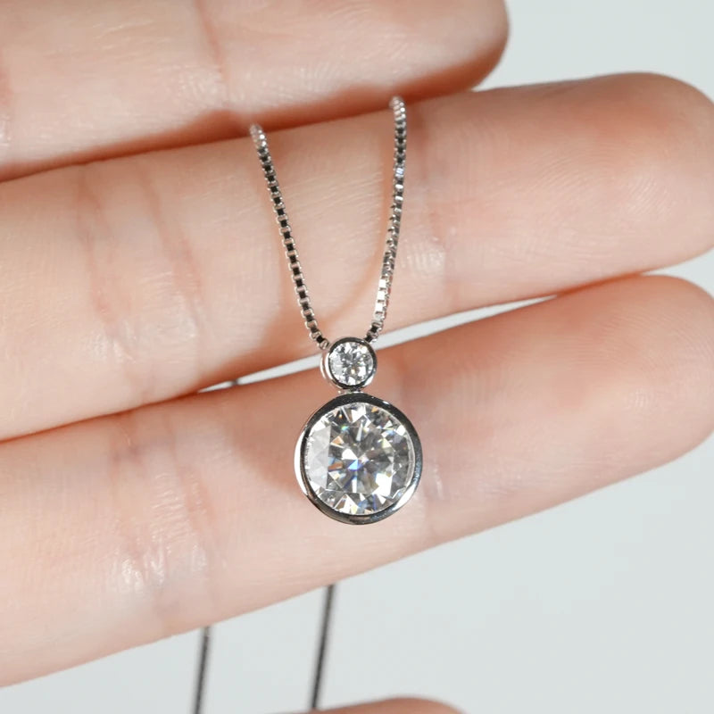 Pendentif serti clos en or blanc et diamants synthétiques montré entre les doigts d'une femme vue de proche.