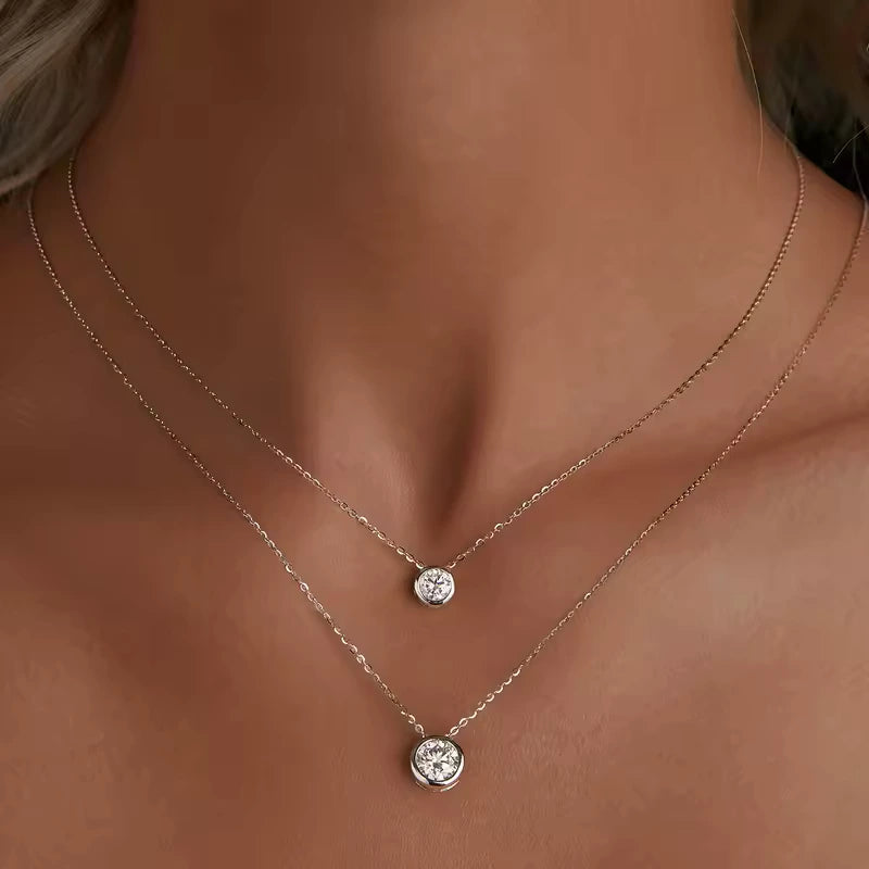 Pendentifs solitaire sertis clos en or blanc raffiné et diamant synthétique 0.5ct et 1ct portés au cou d'une femme vue de face.